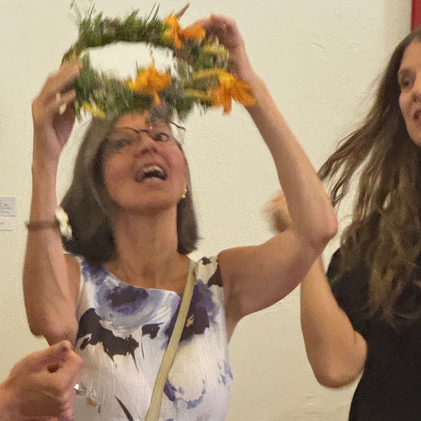 A woman wearing glasses and a floral dress holds a wreath of yellow flowers above her head. Another woman stands behind her, watching.