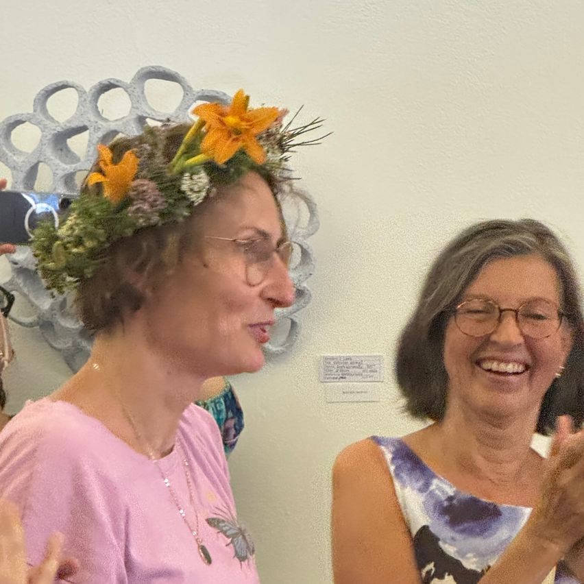 Two women wearing glasses smile at the camera. The woman on the left wears a floral crown on her head. The wall behind them has a hanging art piece and a plaque.