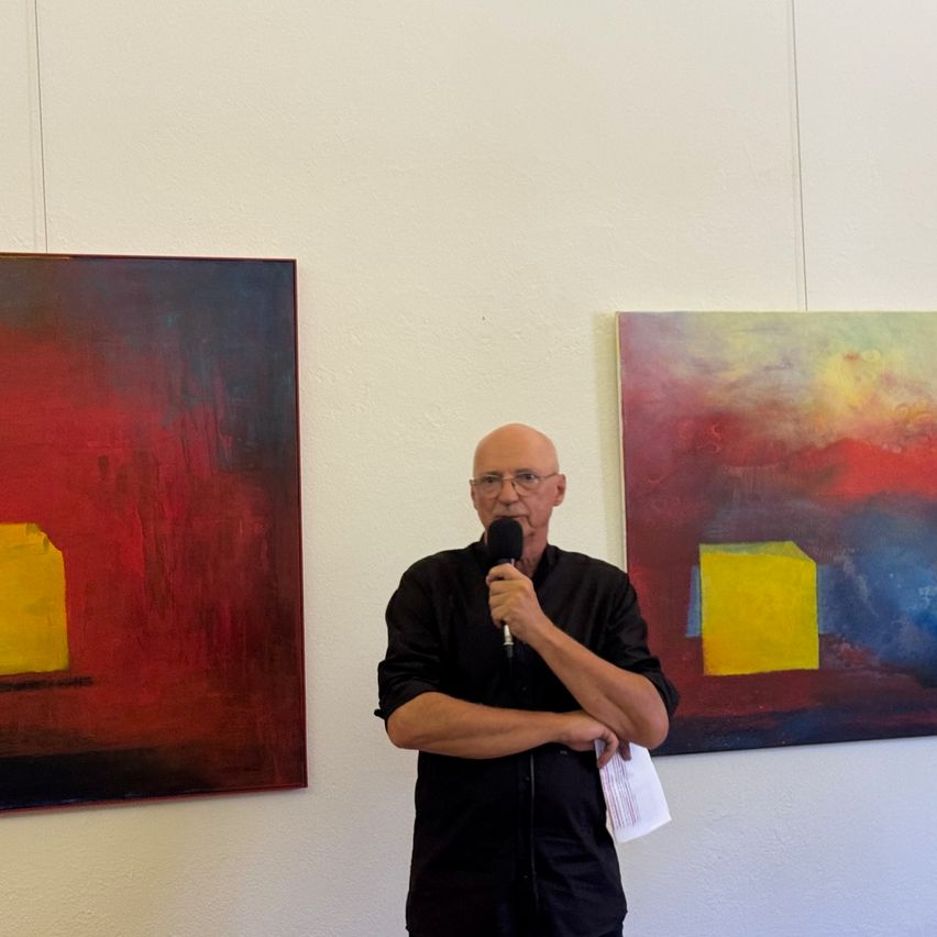 A man in a black shirt speaks into a microphone, standing in front of two abstract paintings on a white wall.