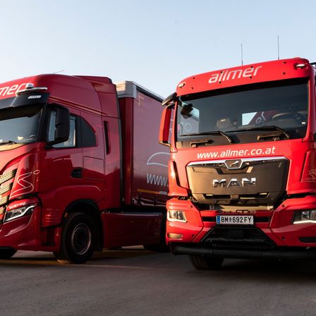 Zwei große rote Lastwagen sind auf einer Betonfläche geparkt. Einer hat 'allmer' vorne und 'www.allmer.co.at' darunter, der andere hat 'www.' an der Seite.