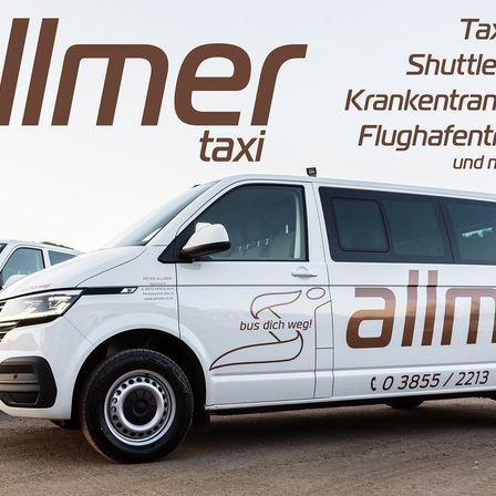 Ein weißer Van von Allmer Taxi ist im Freien geparkt. Der Van hat braune Logos und Texte auf der Seite und der Front, einschließlich einer Telefonnummer.