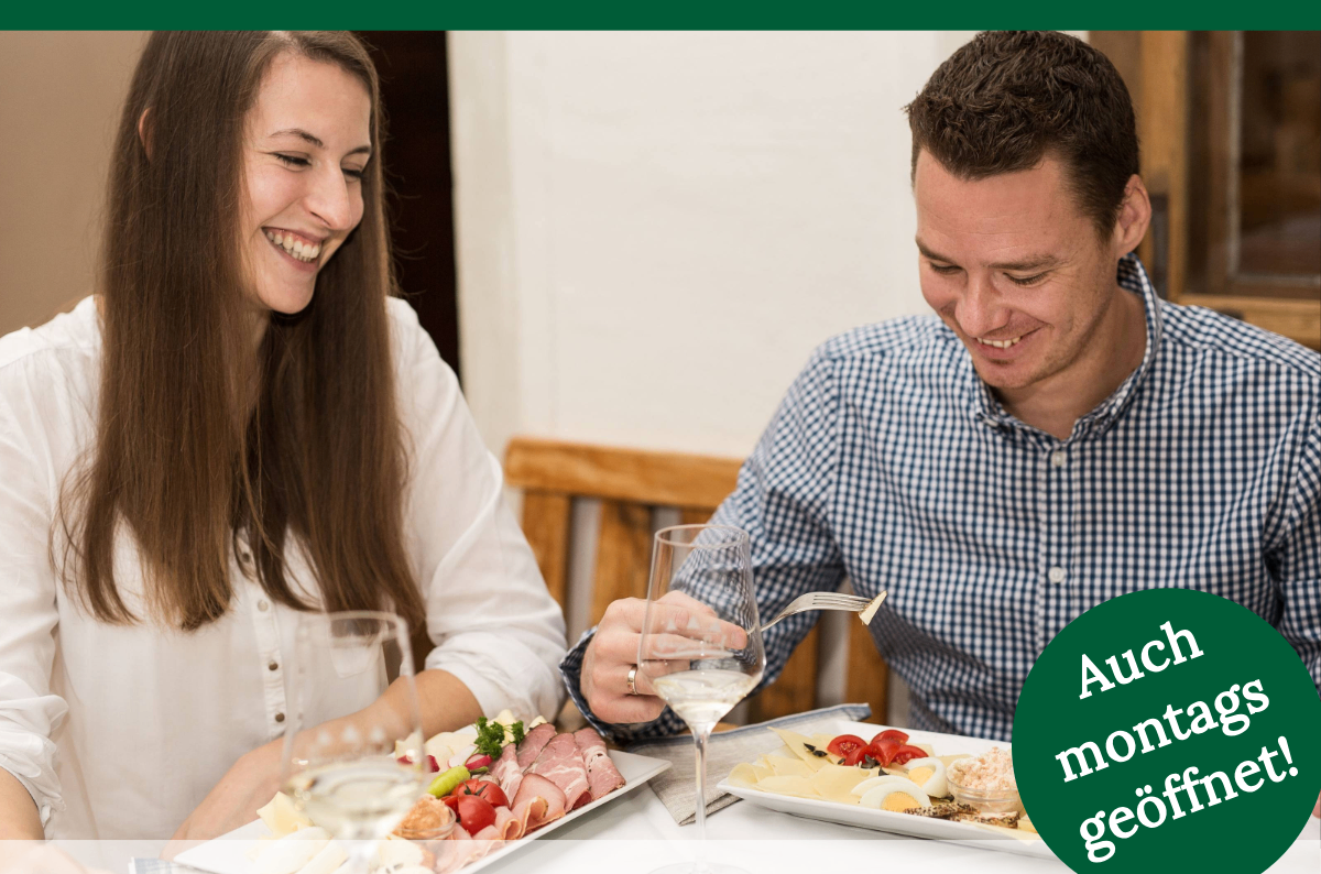Ein Mann und eine Frau lächeln und essen. Der Mann hält eine Gabel und ein Glas Wein. Auf dem Tisch stehen Teller mit Käse, Eiern und Tomaten.