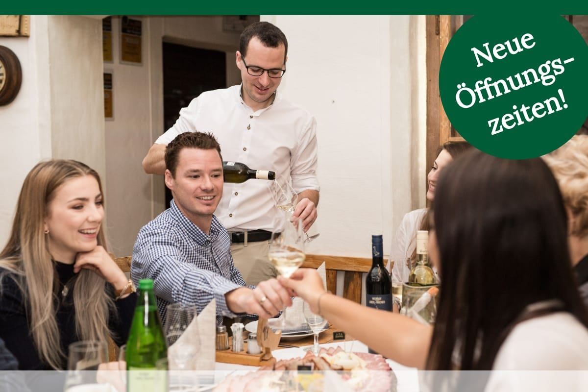 Ein Mann schenkt Wein aus einer Flasche in ein Glas, während eine Gruppe von Leuten um einen Tisch sitzt.