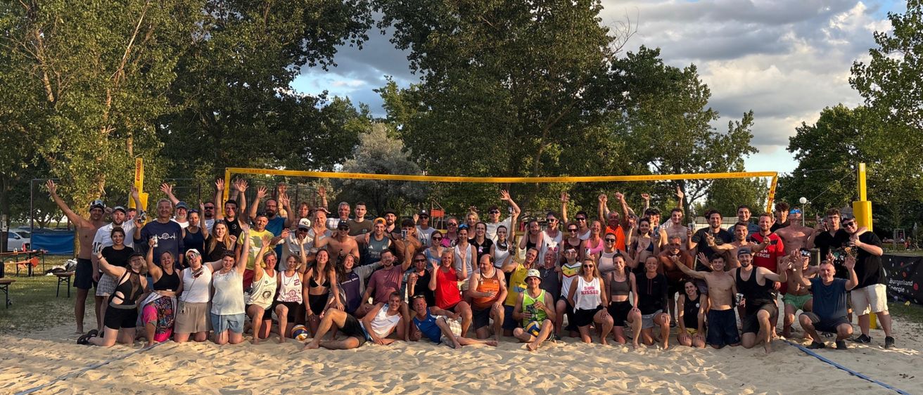 Eine Gruppenaufnahme von Menschen in Strandkleidung posiert vor einem Volleyballnetz auf einem sandigen Strand unter einem teilweise bewölkten Himmel.