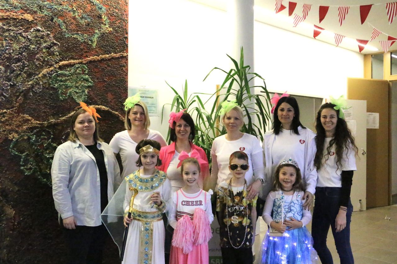 Eine Gruppe von Frauen und Kindern in Kostümen steht in einem Raum. Einige tragen rosa und weiße Kleider, während andere verschiedene thematische Outfits tragen. Sie stehen um eine Pflanze und ein großes Kunstwerk an der Wand herum.