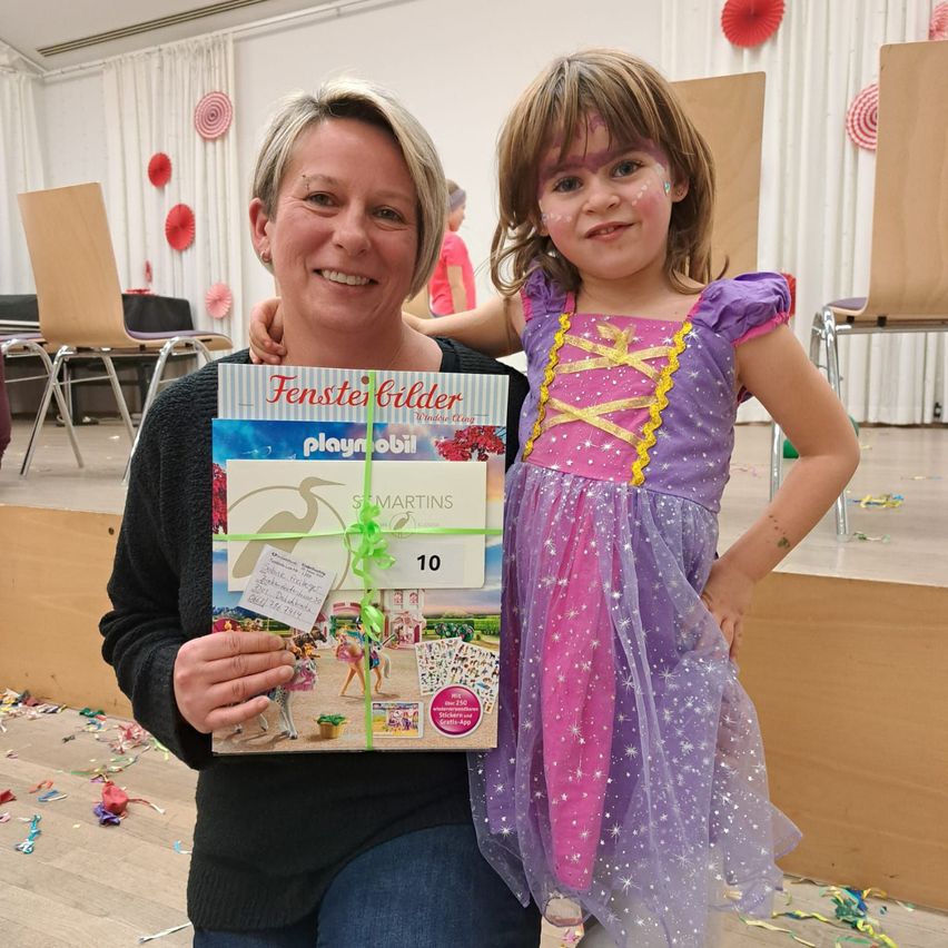 Eine Frau hält ein verpacktes Geschenk in den Händen und lächelt, neben einem kleinen Mädchen in einem Feenkleid. Hinter ihnen ein festlicher Hintergrund mit roten Papierdekorationen.