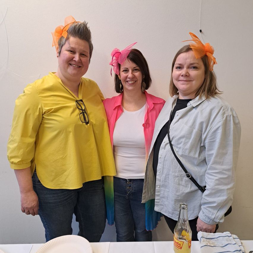 Drei Frauen in Freizeitkleidung mit orangefarbenen Haarbändern stehen nebeneinander vor einer weißen Wand, lächeln und posieren für ein Foto.