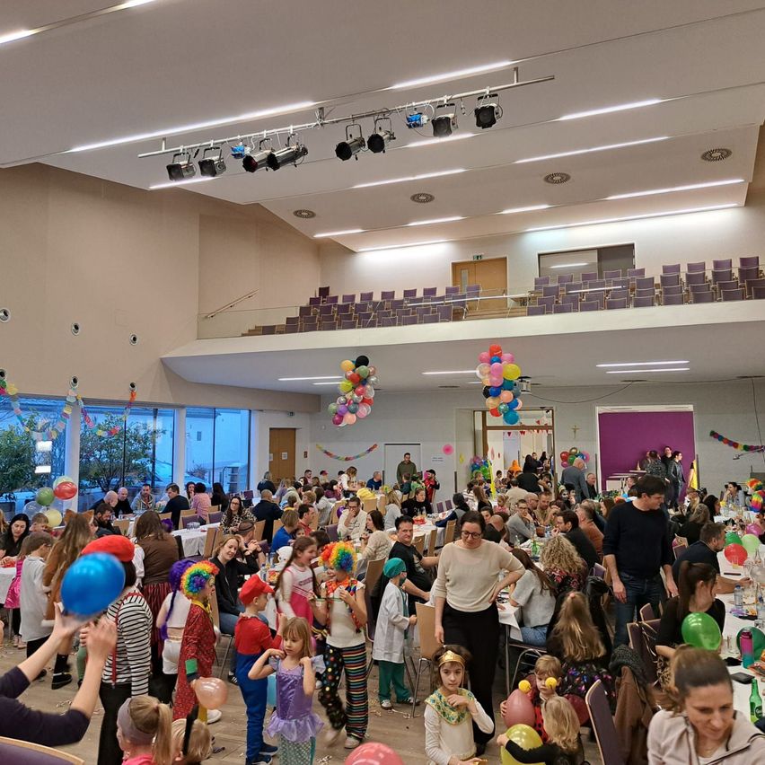 Eine große Menschenmenge versammelt sich drinnen zu einer festlichen Veranstaltung. Bunte Luftballons und Dekorationen schmücken den Raum. Die Menschen sitzen an Tischen, einige stehen und unterhalten sich. Einige Personen halten Luftballons.