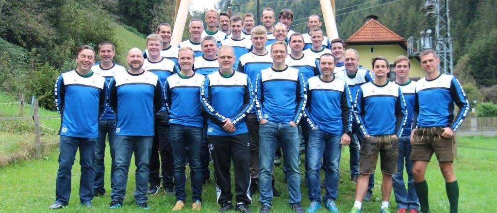 Eine Gruppe von Männern in blauen und weißen Trikots posiert für ein Foto auf einer Rasenfläche mit einer Holzstruktur und Häusern im Hintergrund.