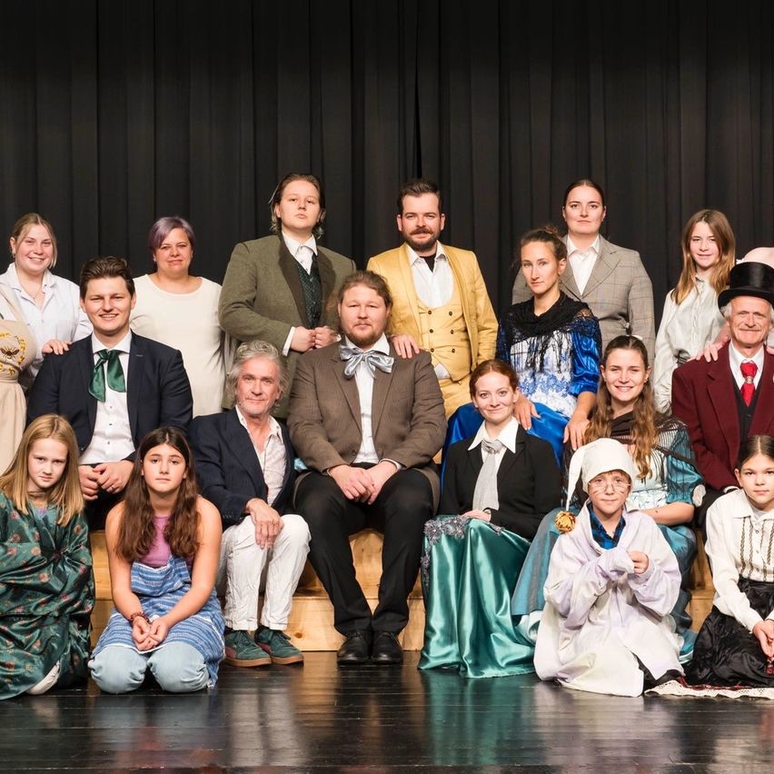 Eine Gruppe von Menschen in Vintage-Kleidung posiert lächelnd auf der Bühne. Sie besteht aus Erwachsenen und Kindern, einige sitzen und andere stehen. Der Hintergrund besteht aus schwarzen Vorhängen.