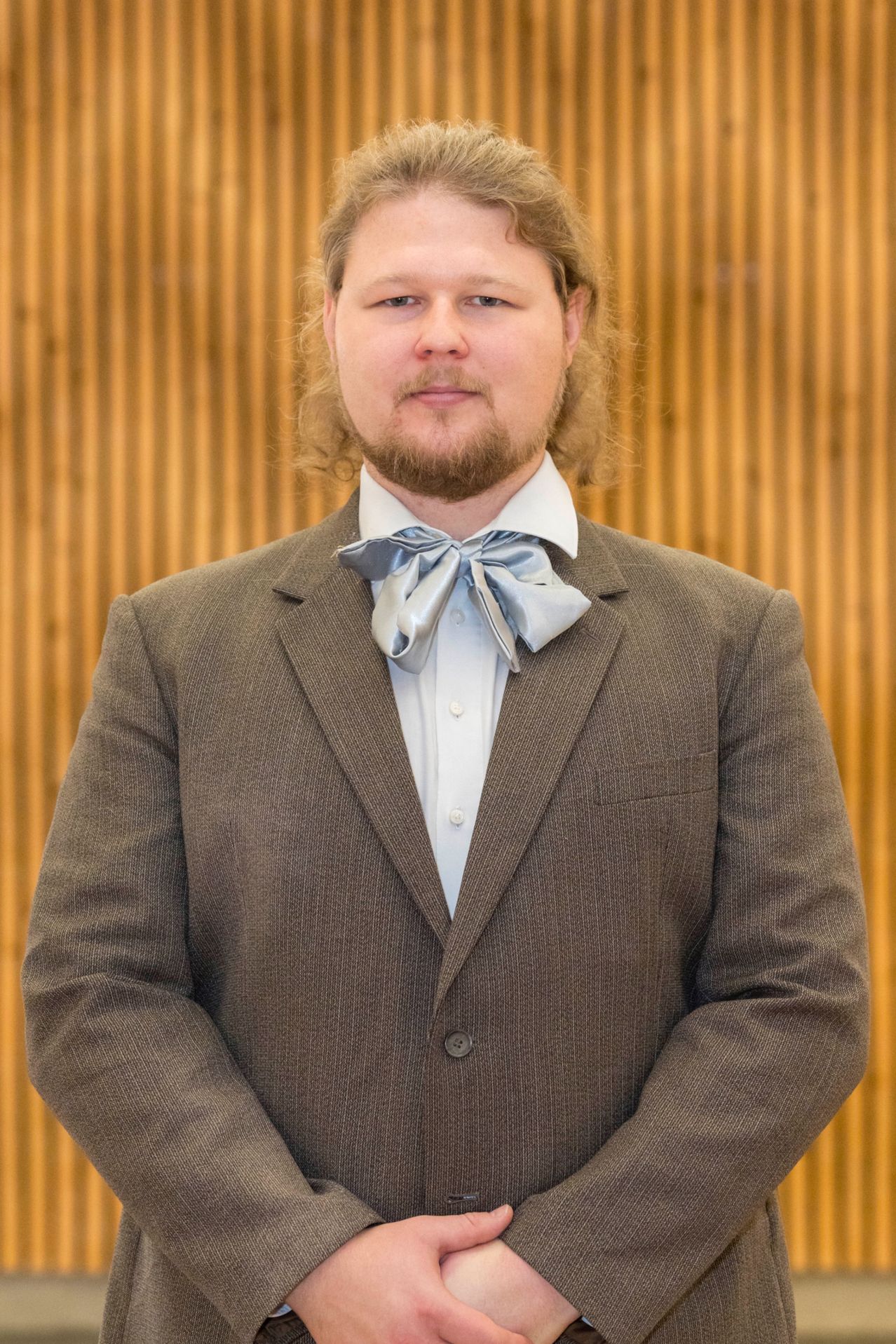 Ein Mann mit langen blonden Haaren und Bart steht vor einer Holzwand, trägt einen braunen Anzug mit weißem Hemd und blauer Fliege.