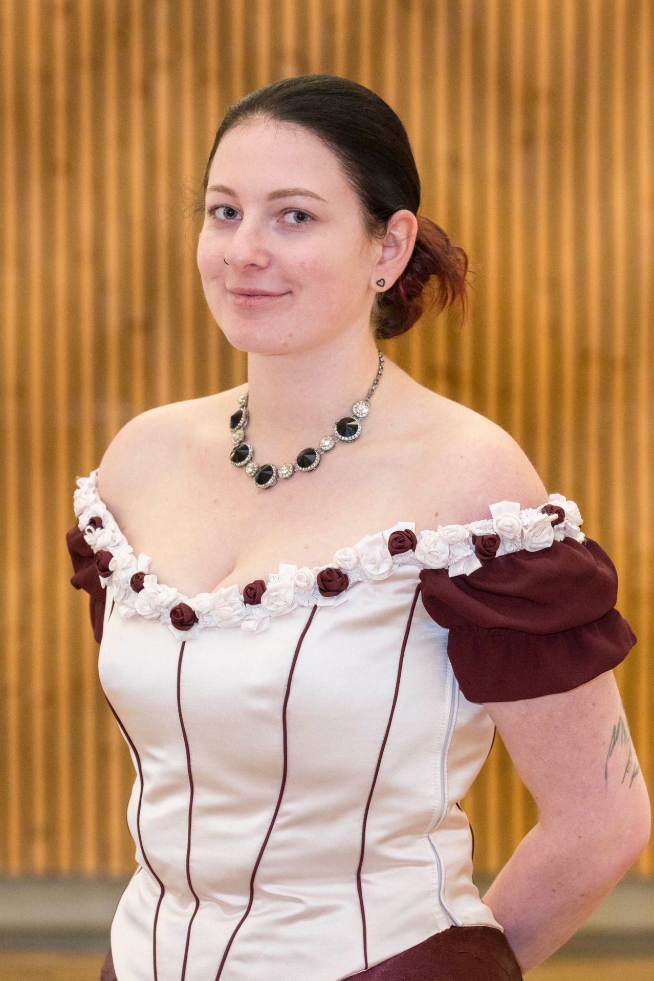 Eine Frau in einem weißen und kastanienbraunen Kleid mit einer Halskette und einem Tattoo am rechten Arm steht vor einer Wand mit einem gestreiften Muster.