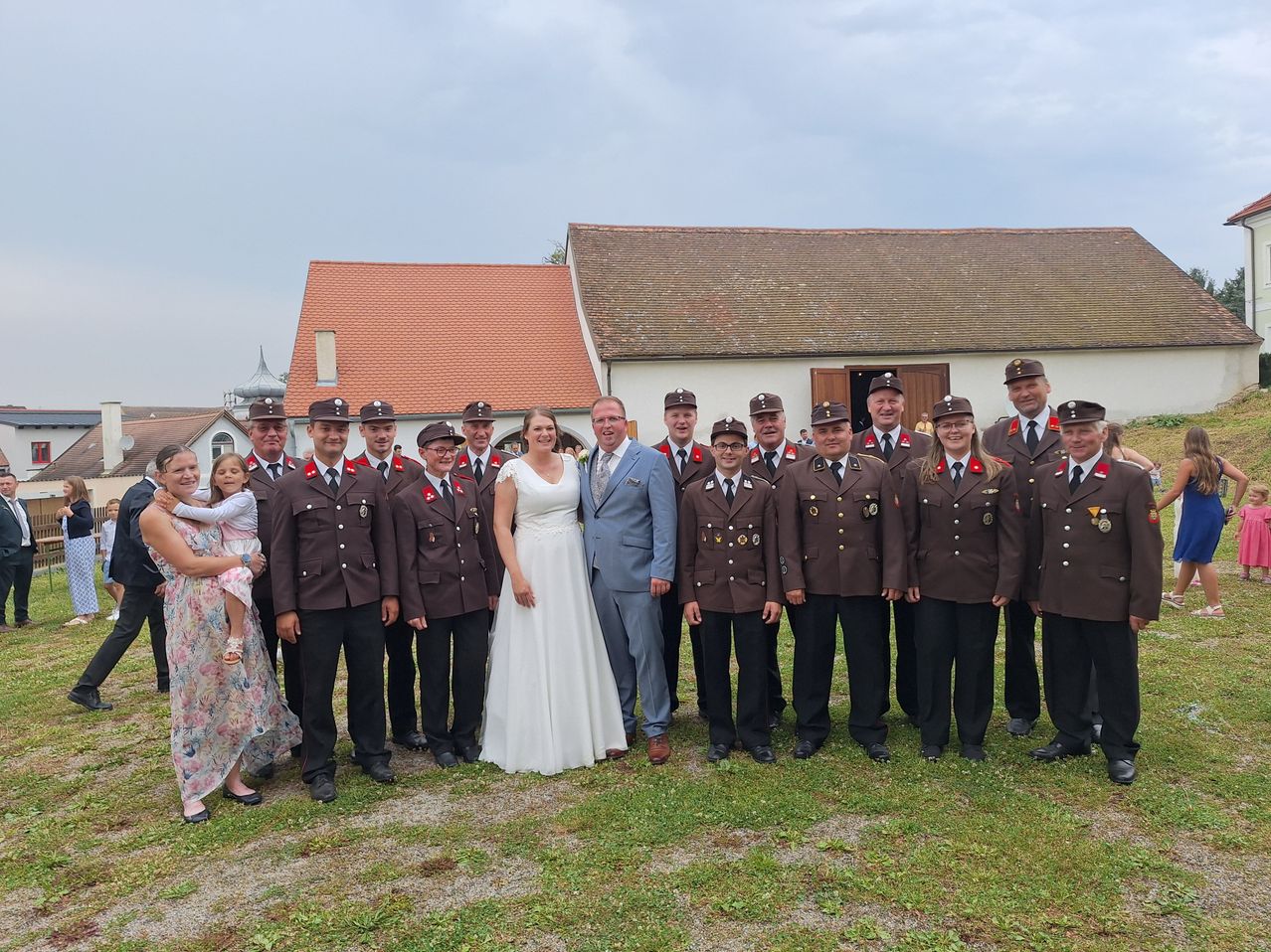 Eine Gruppe uniformierter Personen und ein Paar stehen vor einem Gebäude. Die Frau im Hochzeitskleid und der Mann im Anzug lächeln in die Kamera. Dahinter stehen weitere uniformierte Personen in Reihen.