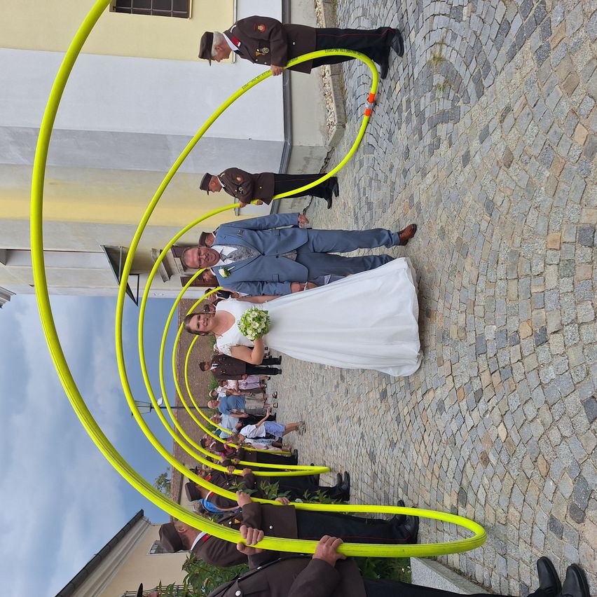 Ein Hochzeitspaar steht unter einem gelben Hula-Hoop-Bogen mit Beamten und Zuschauern auf einem Kopfsteinpflaster unter blauem Himmel.