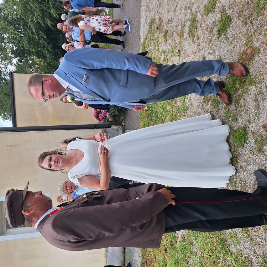 Eine Hochzeitszeremonie im Freien, mit einem Mann und einer Frau in formeller Kleidung. Hinter ihnen stehen mehrere Personen, einige tragen Hüte.