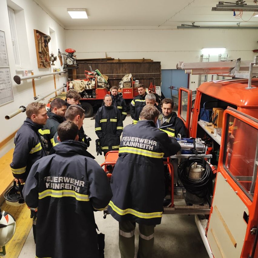 Eine Gruppe von Feuerwehrleuten in Uniform ist in einer Garage um einen Feuerwehrwagen versammelt. Sie diskutieren etwas, während sie um die offene Tür des Trucks stehen.