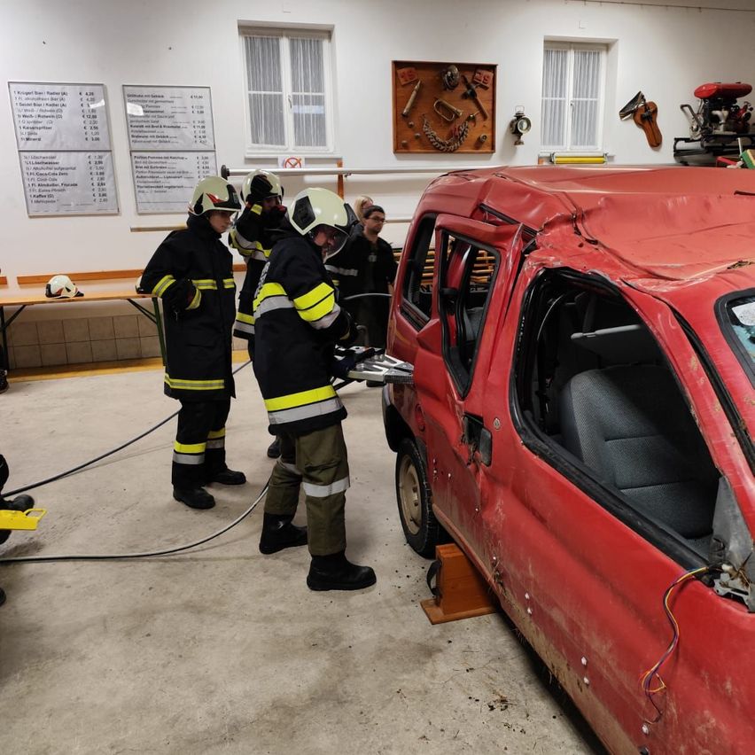Feuerwehrleute arbeiten an einem beschädigten roten Auto mit einem beschädigten Dach und offenen Türen in einer Garage mit Werkzeug und Ausrüstung an der Wand.