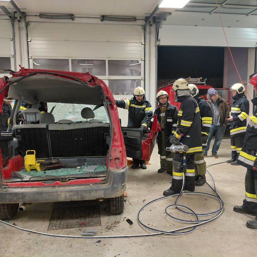 Eine Gruppe von Feuerwehrleuten versammelt sich um ein beschädigtes Fahrzeug in einer Garage. Sie tragen Helme und Uniformen. Ein Feuerwehrmann hält ein Werkzeug in der Hand. Die hintere Tür des Fahrzeugs ist offen und die Scheibe ist zerbrochen.