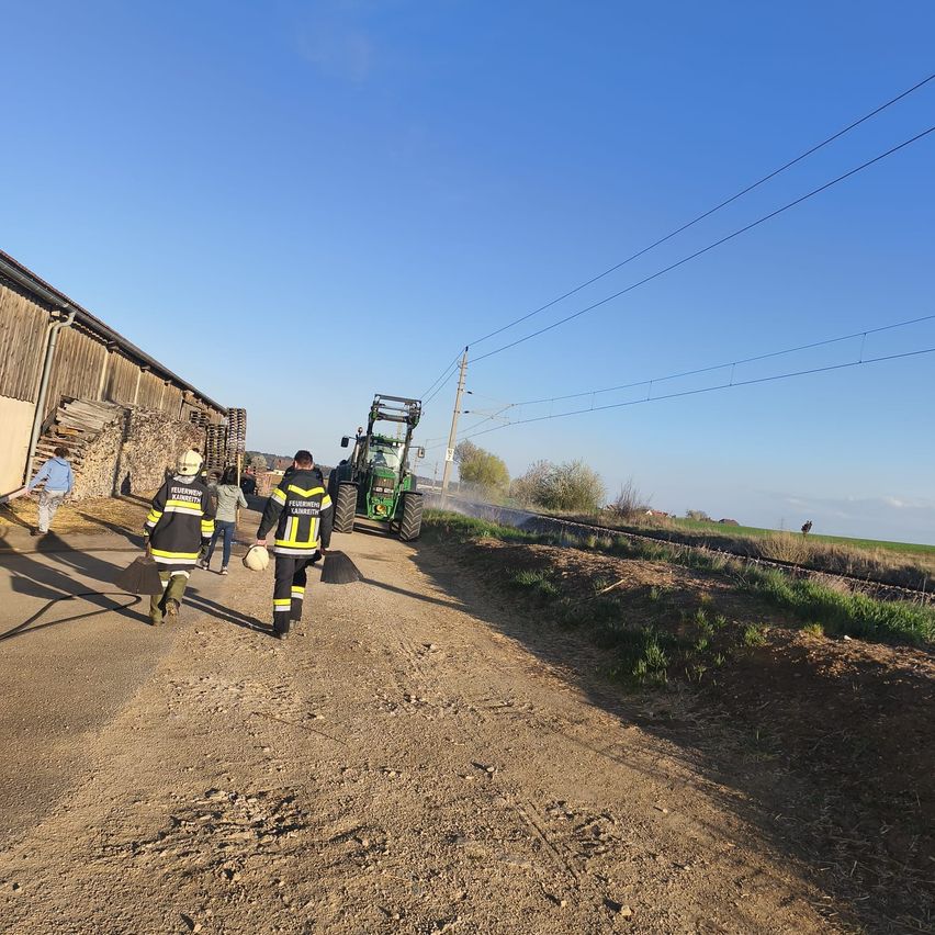 Zwei Feuerwehrleute gehen einen Feldweg entlang, einer trägt einen Helm und einen Feuerlöscher. Ein Traktor ist auf der Bahn, und Stromleitungen verlaufen rechts.