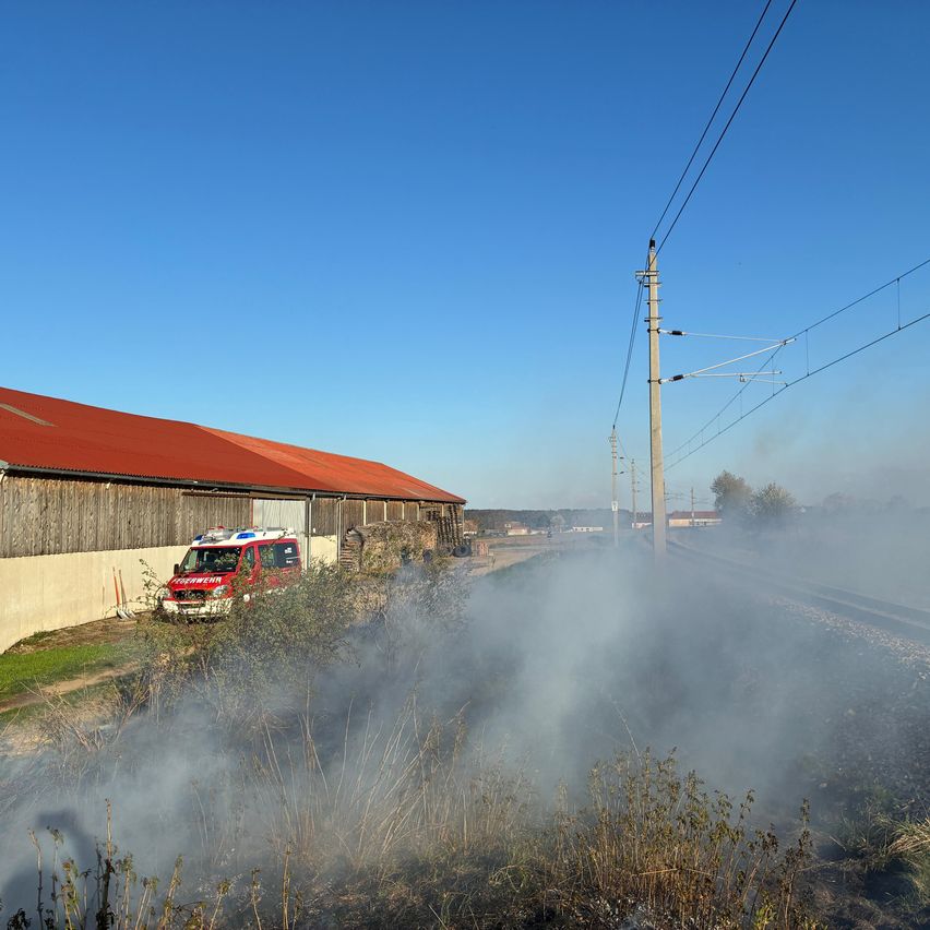 Ein Feuerwehrauto steht neben einem Gebäude mit rotem Dach. Rauch steigt vom Boden auf, in der Nähe einer Stromleitung und einer Bahnlinie.