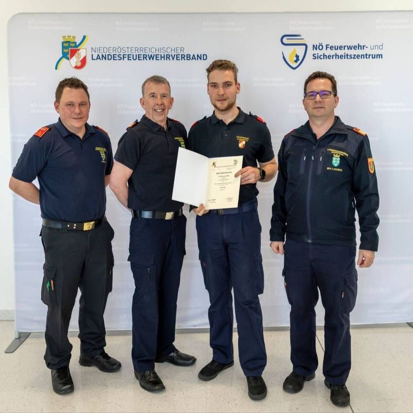 Vier Männer in Uniform stehen in einer Reihe und lächeln. Einer hält ein Zertifikat, und der Hintergrund zeigt Logos und Text.