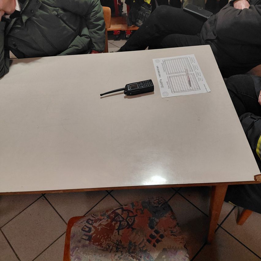 Ein Tisch mit einem Handy und einem Papierblatt darauf. Drei Personen sitzen darum. Der Boden ist gefliest, und ein Stuhl steht vor dem Tisch.