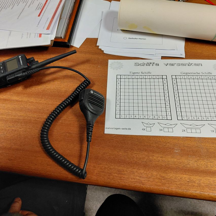 Ein Schreibtisch mit einem schwarzen Motorola-Funkgerät, gewickeltem Kabel und einem Papier mit einem Schlachtschiffspiel. Andere Papiere und ein gerolltes Papier sind auf dem Schreibtisch.