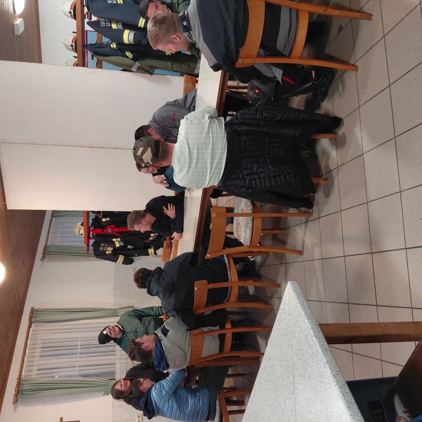 Eine Gruppe von Feuerwehrleuten versammelt sich um einen Tisch in einem Raum. Einige stehen, andere sitzen und tragen ihre Uniformen. Der Raum hat weiße Wände und geflieste Böden. Links befinden sich Fenster mit Vorhängen.