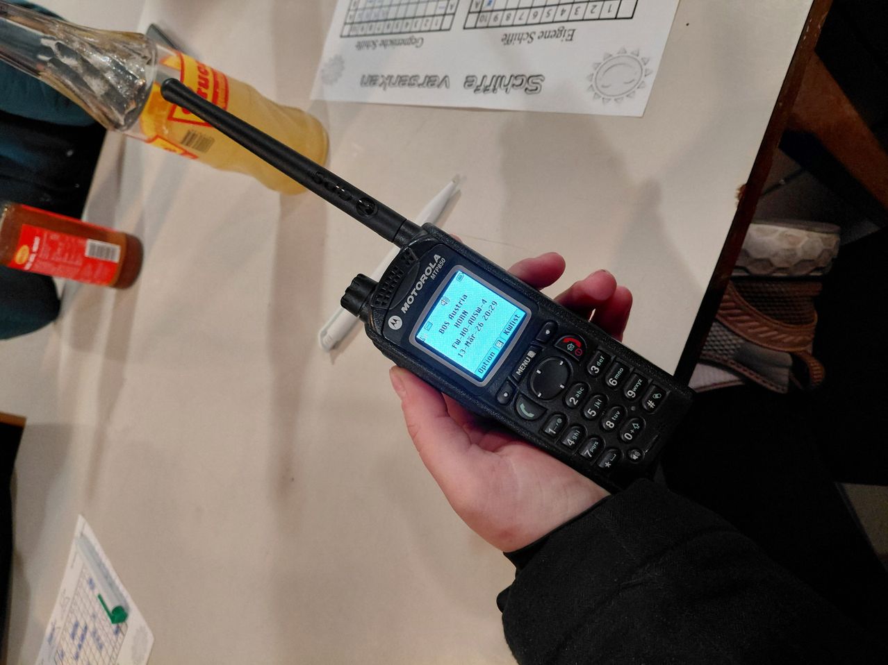 Ein Motorola-Funkgerät wird in einer Hand gehalten, mit einem blauen Bildschirm, der 'Fr-Mon' anzeigt. Dahinter befindet sich ein Tisch mit verschiedenen Gegenständen, darunter eine Flasche, ein Marker und ein Stück Papier.