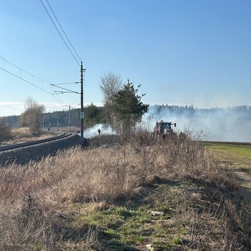 Ein Traktor spritzt Wasser auf ein Feuer in der Nähe einer Eisenbahnlinie mit Stromleitungen, unter einem klaren Himmel.