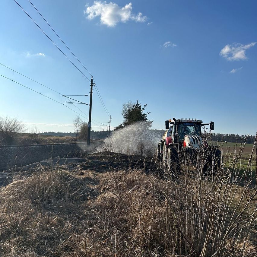 Ein Traktor sprüht Wasser auf ein Grasfeld mit einer Stromleitung und einer Bahn im Hintergrund. Der Himmel ist blau mit einigen Wolken.