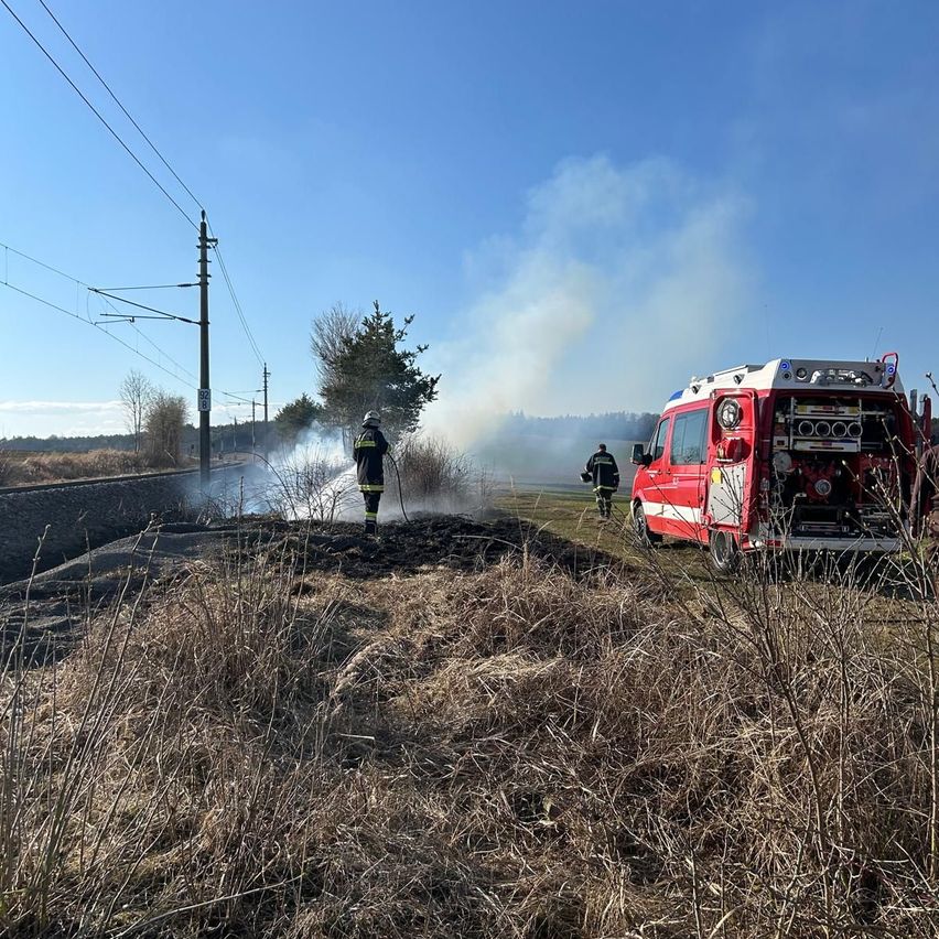 Feuerwehrleute löschen ein Feuer auf einer Bahnstrecke, mit aufsteigendem Rauch und einem roten LKW in der Nähe.