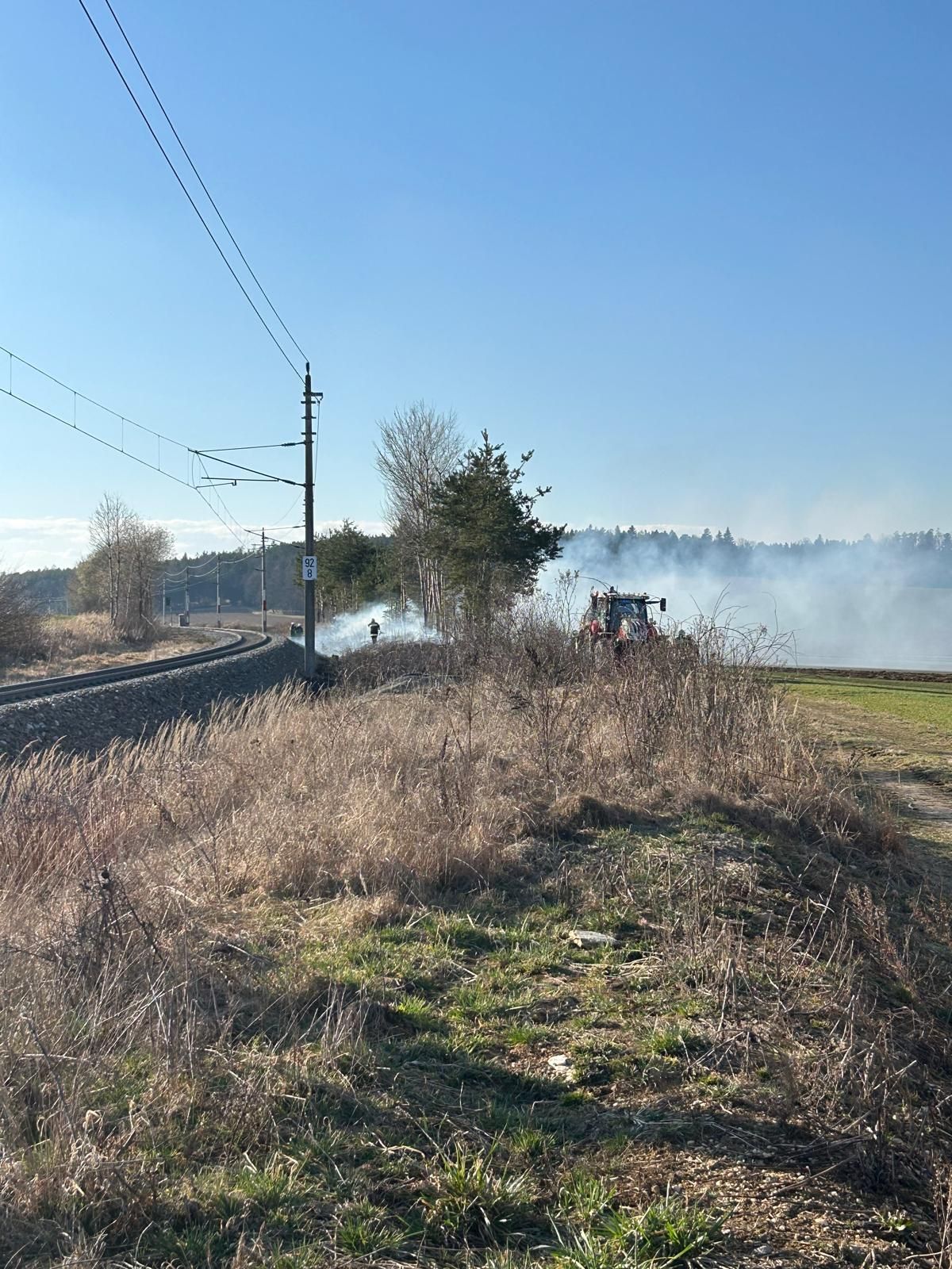 Ein Feld mit hohem Gras in der Nähe einer Bahnstrecke, ein Traktor spritzt Wasser, um ein Feuer zu löschen, und Rauch steigt vom Boden auf.