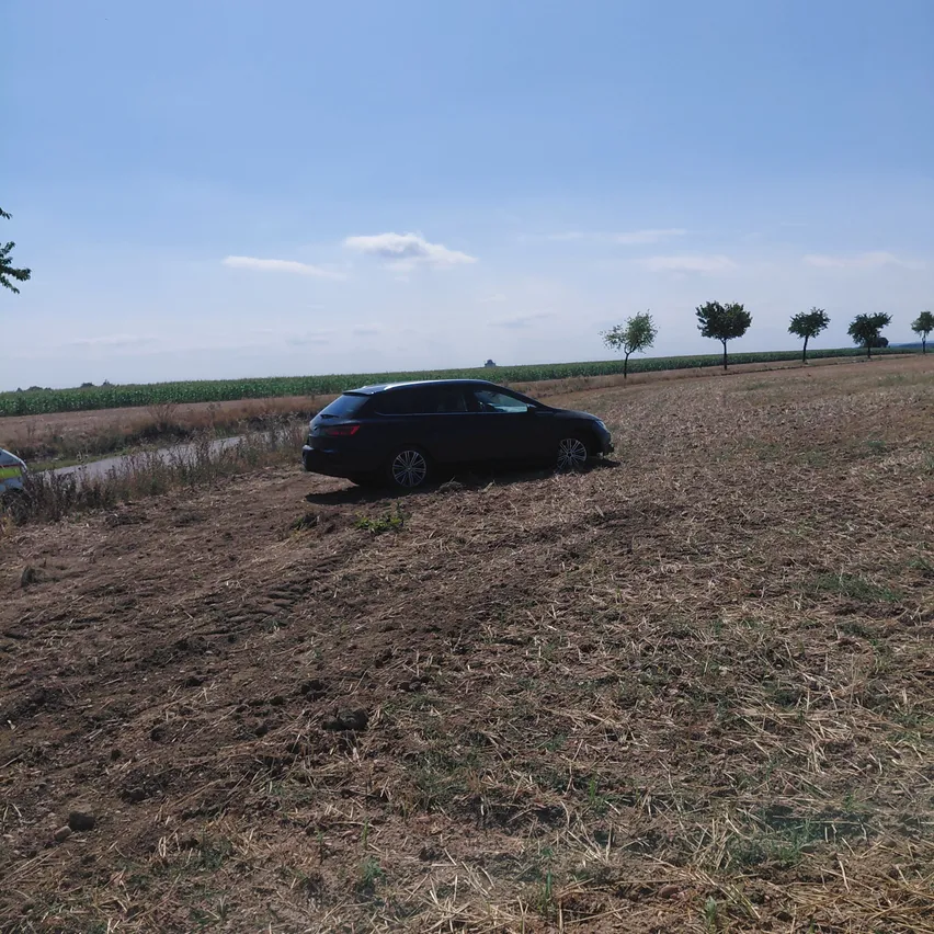Ein schwarzes Auto steckt in einem Feld mit trockenem Gras und kleinen Bäumen im Hintergrund.