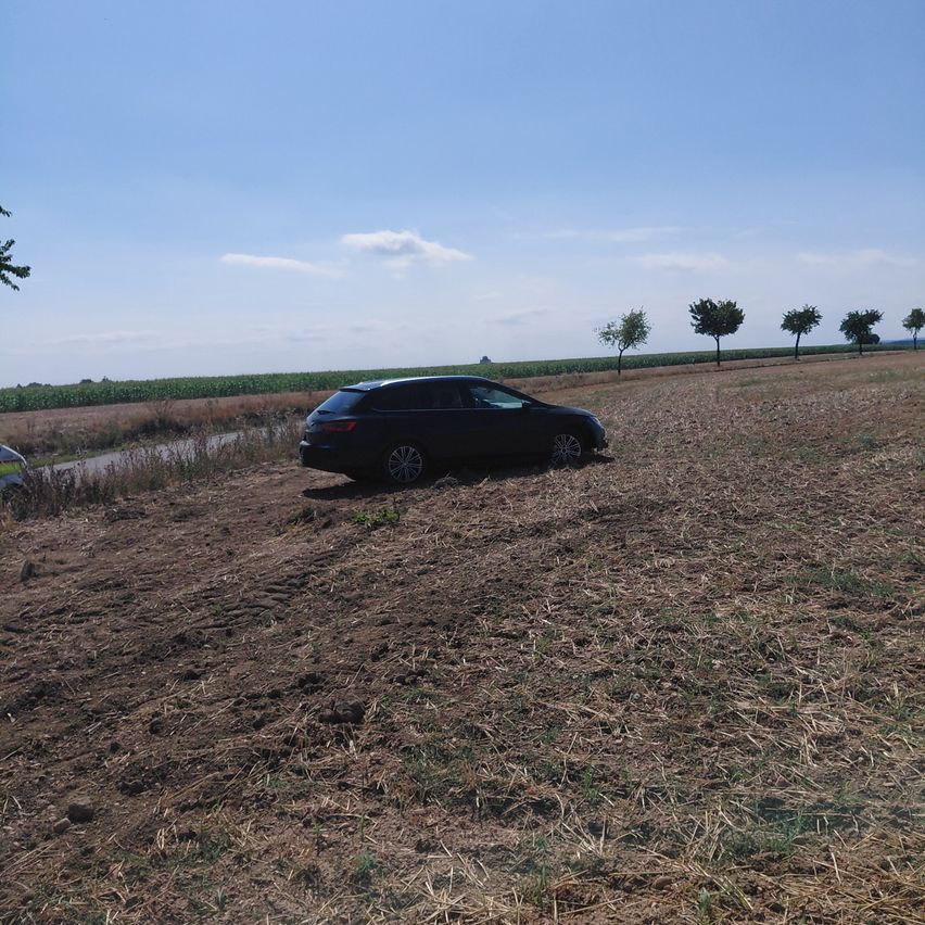 Ein schwarzes Auto steckt in einem Feld mit trockenem Gras und kleinen Bäumen im Hintergrund.