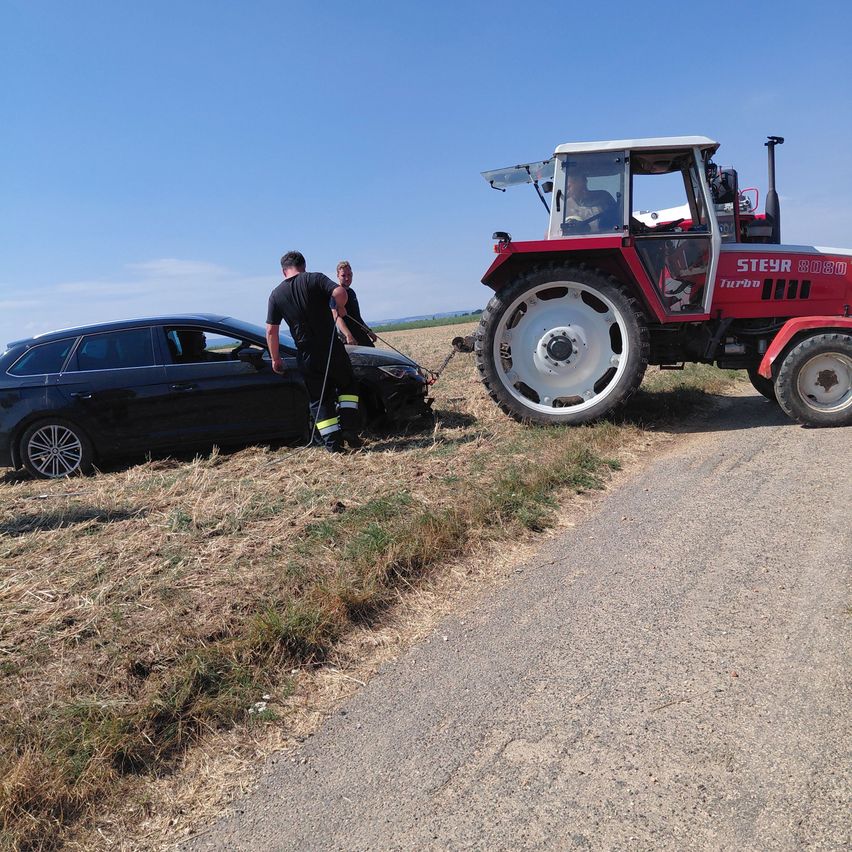 Ein roter Traktor zieht ein schwarzes Auto mit einem Seil von der Straße. Zwei Personen stehen neben dem Auto.