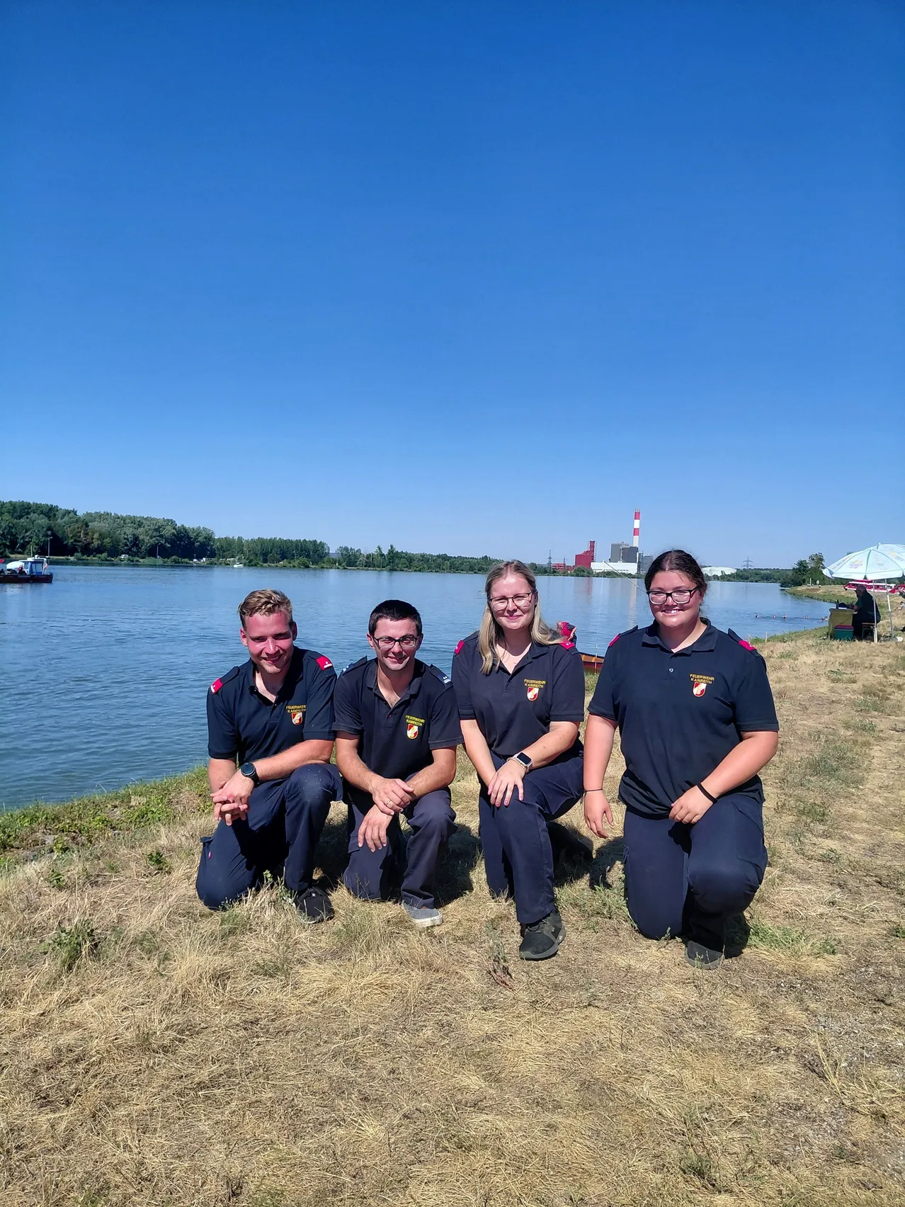 Vier Personen in Uniform posieren für ein Foto am See mit klarem blauem Himmel und entfernten Bäumen.