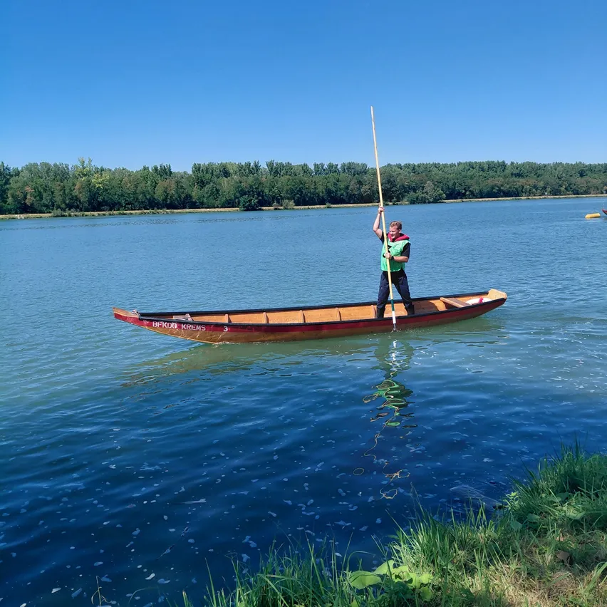 Eine Person steht in einem roten Kanu auf einem See, hält ein Paddel und hat einen klaren blauen Himmel und Bäume im Hintergrund.