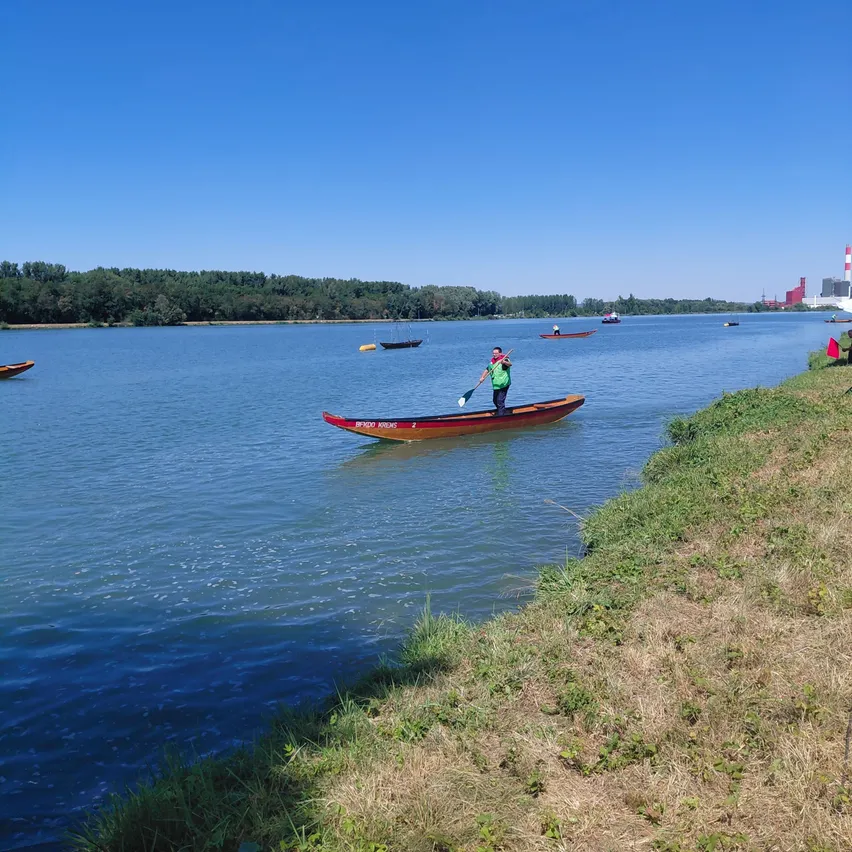 Eine Person in einem grünen Shirt paddelt ein rotes Kajak auf einem ruhigen See mit anderen Booten und einem Ufer im Hintergrund.