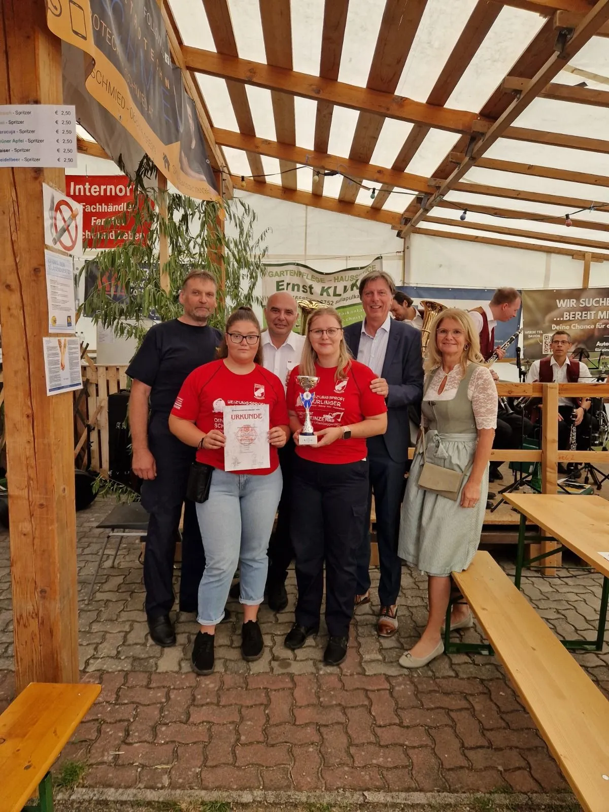Fünf Personen stehen in einer Gruppe, zwei Frauen halten Trophäen und ein Mann steht hinter ihnen. Sie alle lächeln und posieren für ein Foto. Hinter ihnen sitzen ein paar Leute auf einer Bank und spielen Musikinstrumente. Einige Tafeln sind an der Wand montiert.