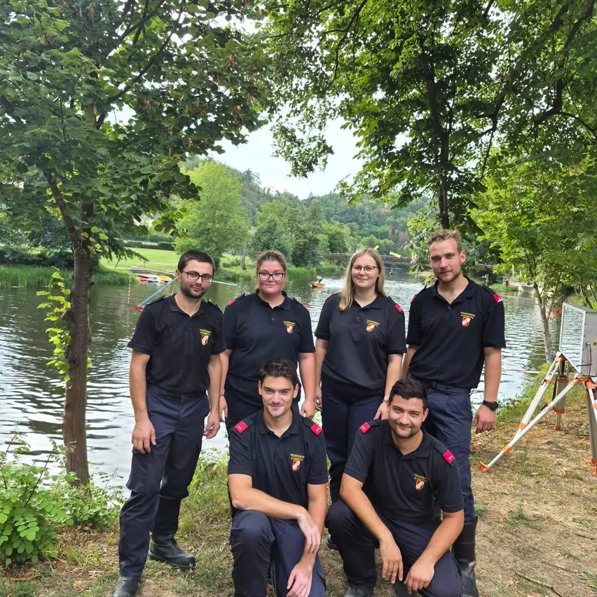Eine Gruppe von Menschen in Uniform posiert für ein Foto am See mit Bäumen und einer Brücke im Hintergrund.