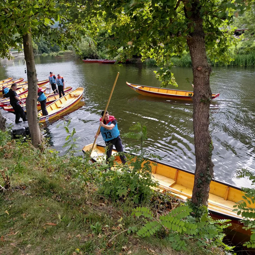 Eine Gruppe von Menschen in blauen Westen und nummerierten Shirts steht am Ufer und bereitet sich darauf vor, gelbe Boote auf einem Fluss zu rudern. Eine Person ist bereits in einem Boot und hält ein Paddel. Bäume und Grünpflanzen säumen das Flussufer.