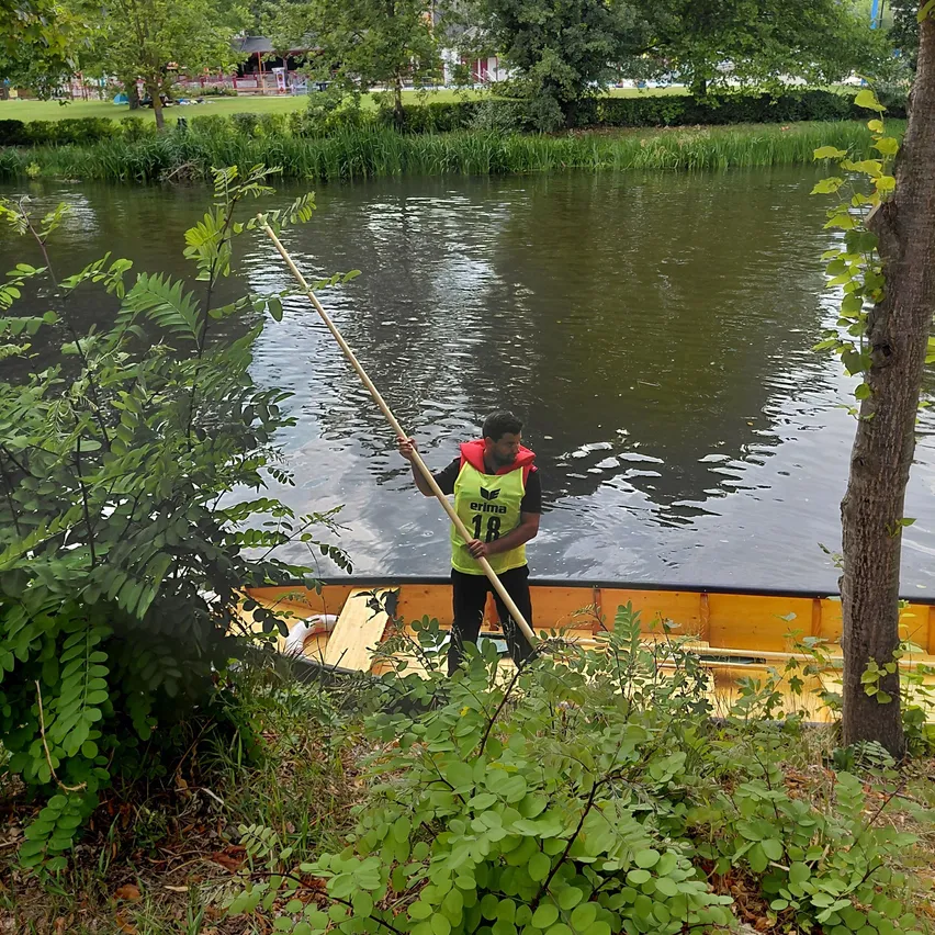 Ein Mann in einem gelben Sicherheitsweste steht in einem gelben Boot, hält einen langen Stab, nahe einem Fluss mit Bäumen und Grün.