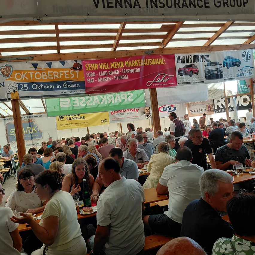 Eine große Menschenmenge sitzt an Tischen in einem Außenbereich, möglicherweise auf einem Festival. Über ihnen hängen Banner und Werbung, darunter eine für die Vienna Insurance Group.