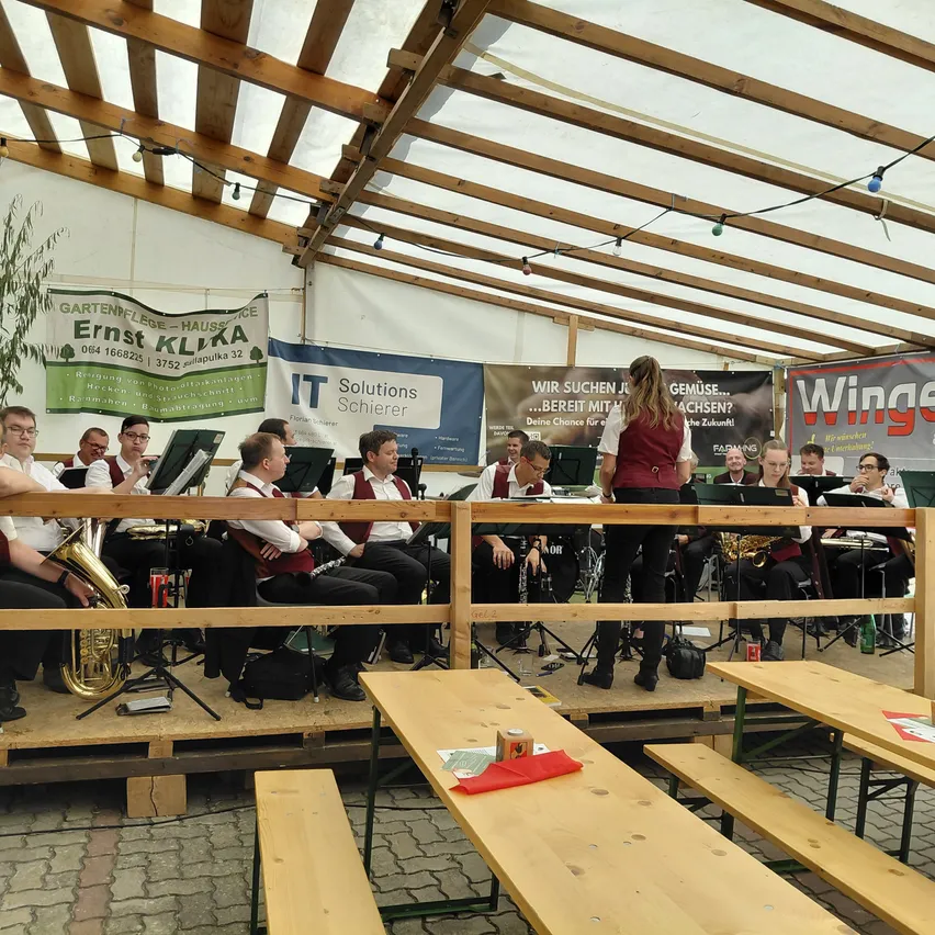 Ein Blasorchester spielt auf einer Bühne unter einem Zelt mit Holzbalken, eine Frau steht davor. Das Publikum sitzt auf Bänken vor der Bühne.