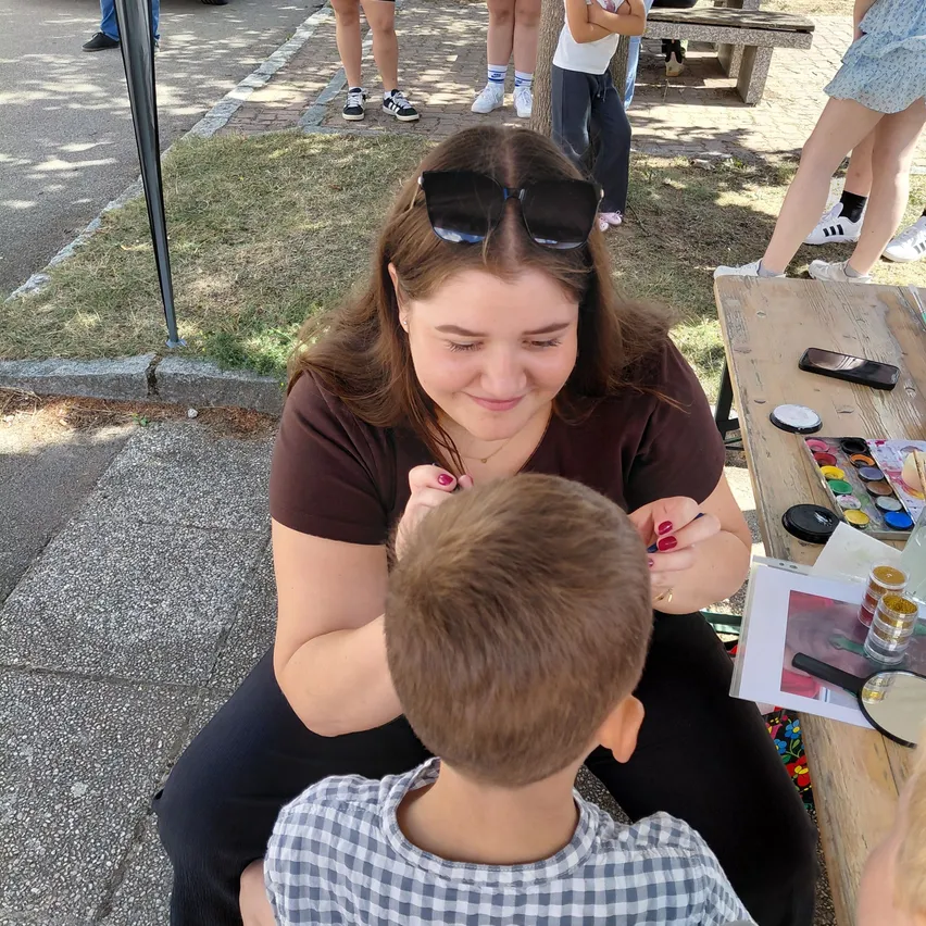 Eine Frau mit Sonnenbrille sitzt auf dem Boden und schminkt das Gesicht eines jungen Jungen. Der Junge trägt ein kariertes Hemd. Dahinter steht ein Holztisch mit Farbe und einem Handy. Im Hintergrund stehen und sitzen Leute auf dem Rasen.