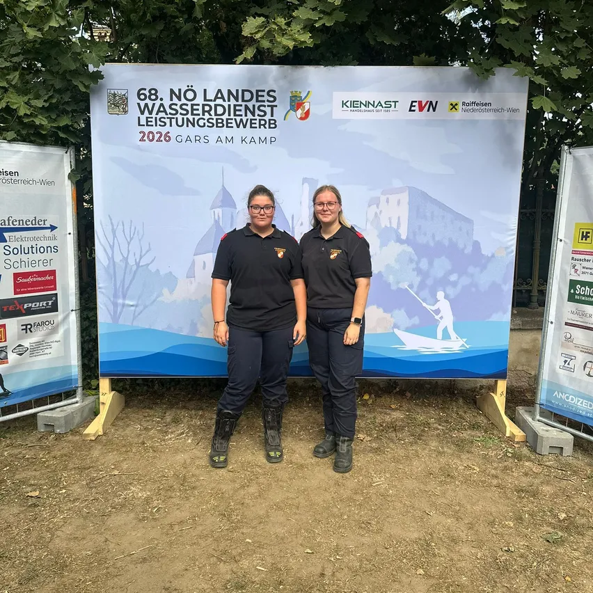 Zwei Frauen in Uniform stehen vor einem großen Banner, das eine Wasserdienstleistungsveranstaltung im Jahr 2026 ankündigt. Sie lächeln und scheinen für ein Foto zu posieren.
