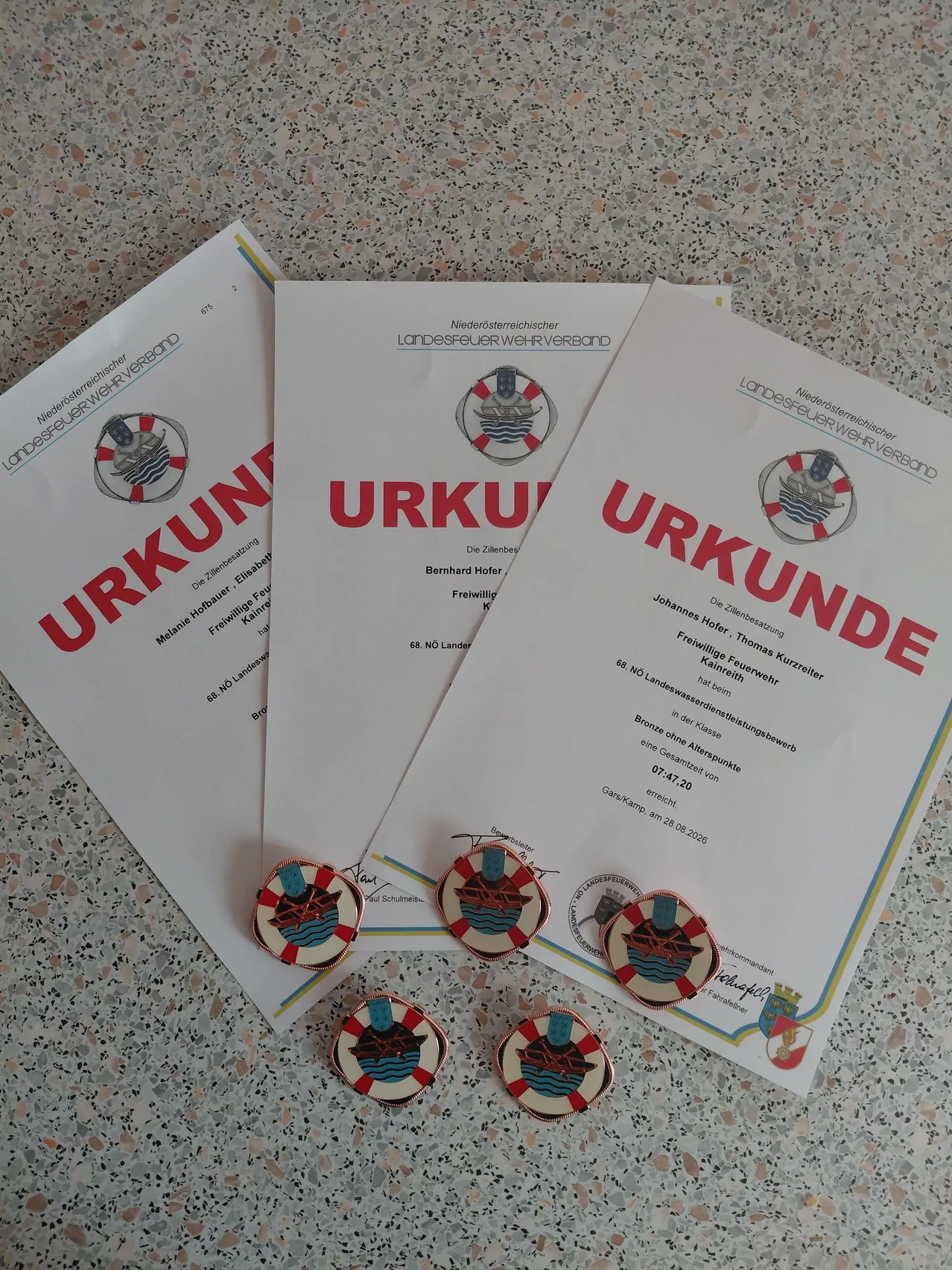 Drei weiße Urkunden mit rotem Text 'URKUNDE' und einem Logo oben, auf einer Marmoroberfläche platziert. Unter den Urkunden befinden sich vier kreisförmige Anstecknadeln mit einem Rettungsring-Logo.