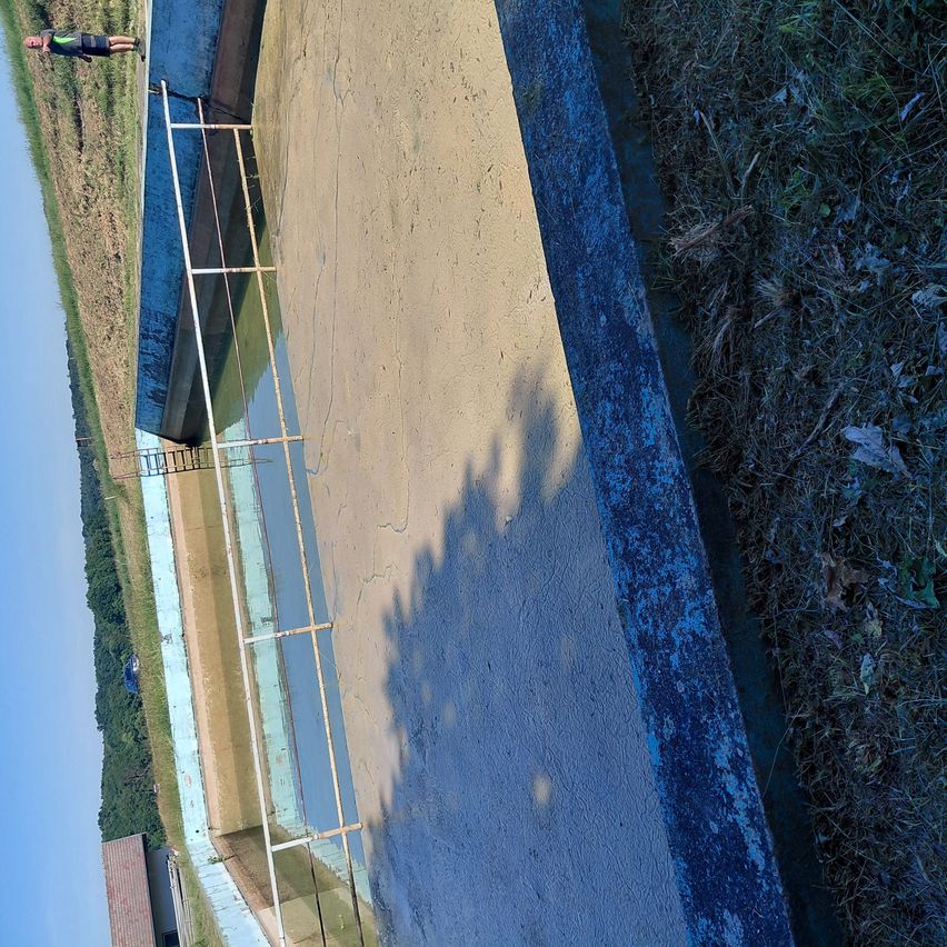 Luftaufnahme eines leeren Schwimmbeckens mit einer Metallleiter an der Seite. Das Becken ist von Gras und Erde umgeben. Der Poolbereich ist nass, und es gibt einen Schatten auf dem Boden.