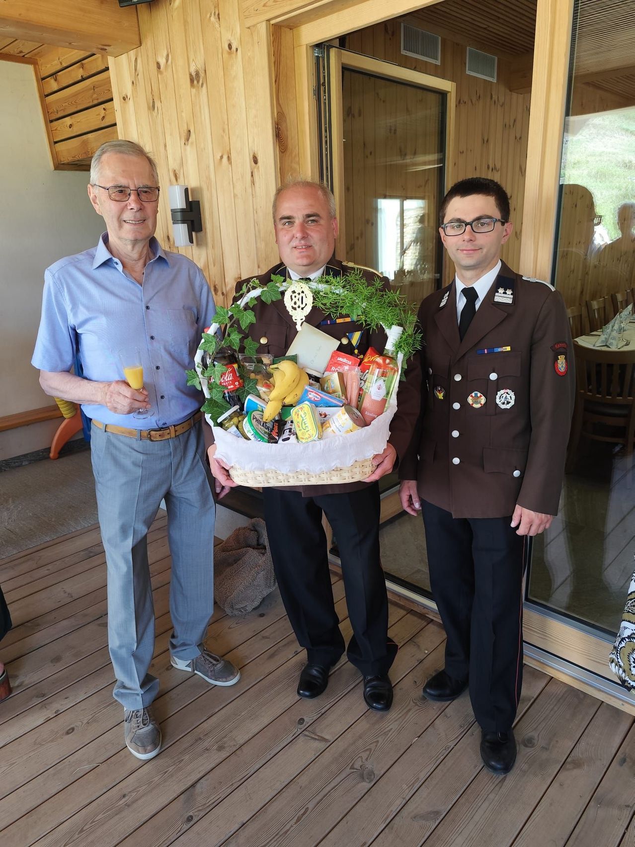 Drei Männer stehen auf einer Veranda. Der Mann in der Mitte hält einen Korb voller Essen und Getränke. Der Mann rechts trägt eine militärische Uniform. Der Mann links trägt einen Gürtel und hält ein Glas Orangensaft.